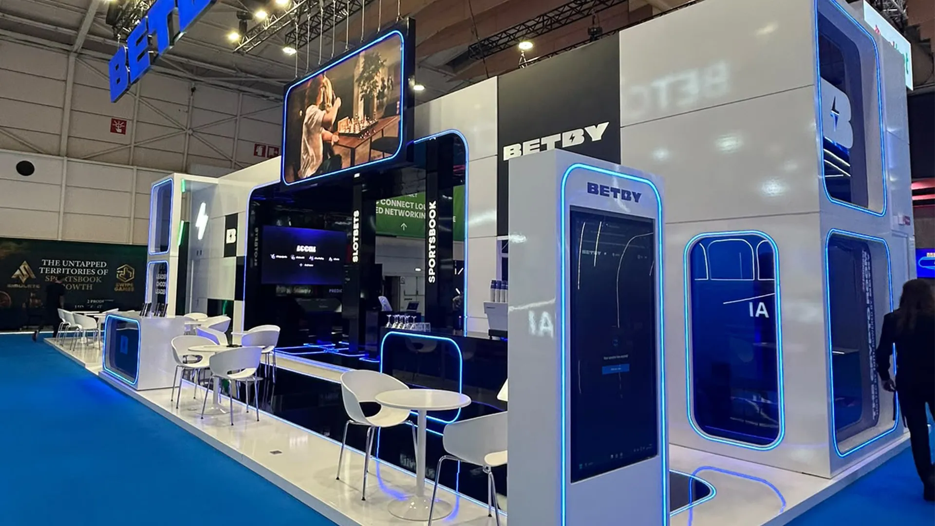 Angled view of BETBY’s SBC Summit Lisbon 2026 stand with suspended screen, blue illuminated edges and open meeting tables.