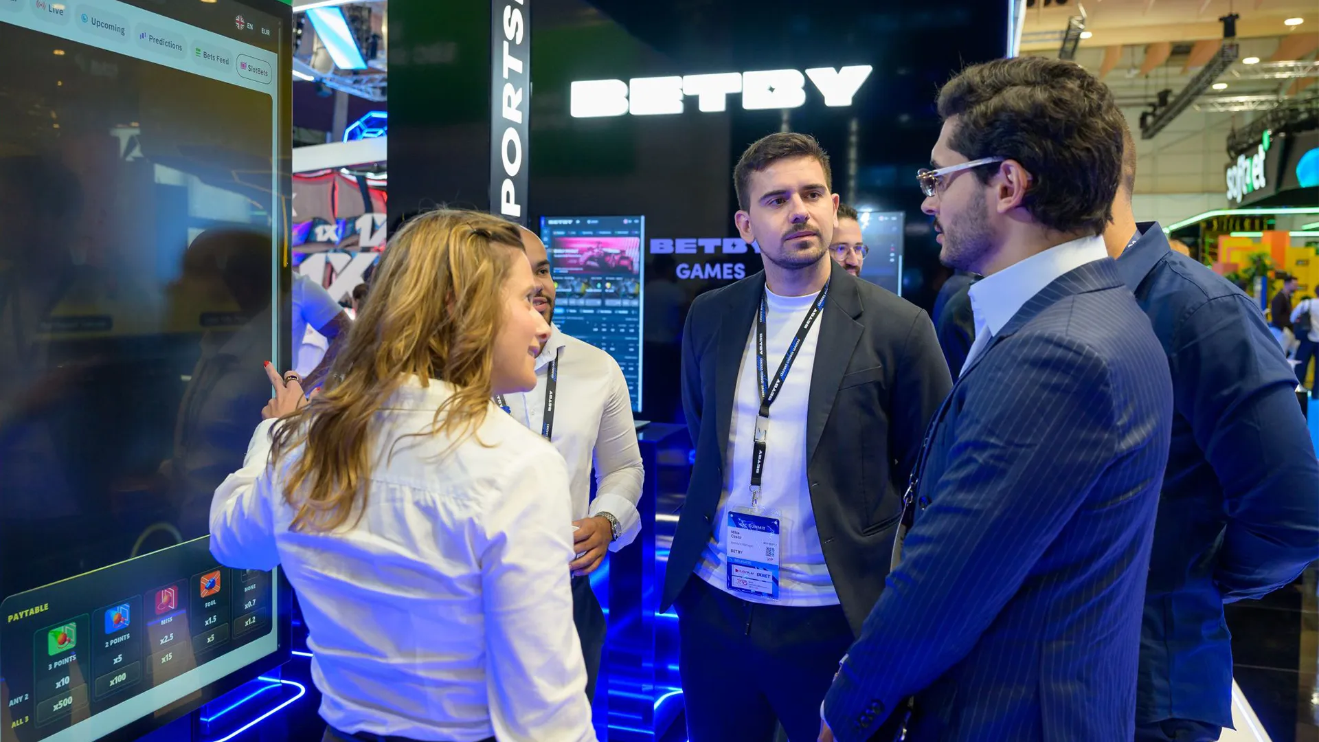 A group gathered beside a large SlotBets display during a product demonstration on the BETBY stand.