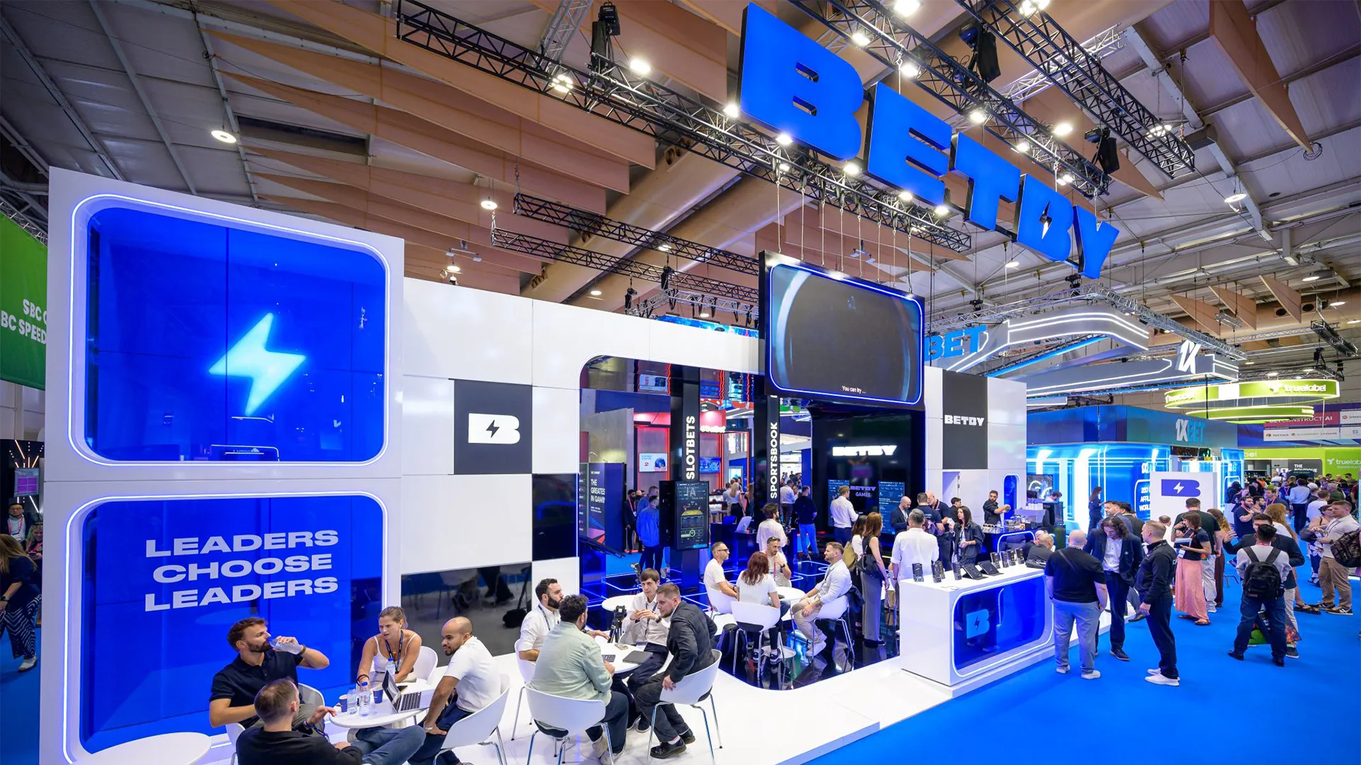 Wide view of the active BETBY stand with overhead branding, suspended screen and visitors seated at meeting tables.