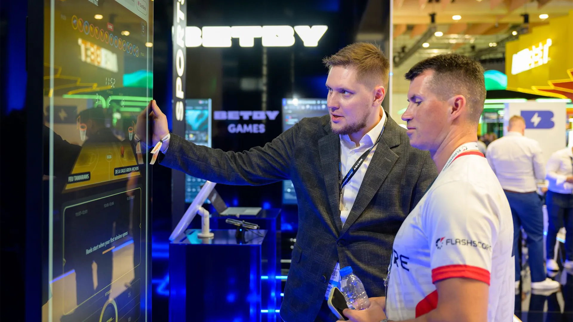 A presenter explaining the SlotBets basketball interface to a visitor on a large vertical screen.