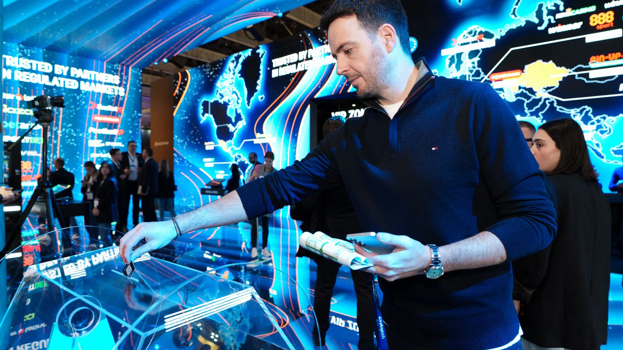 Visitor places a chip on a lit interactive table at the Digitain stand, with large digital world map graphics behind.