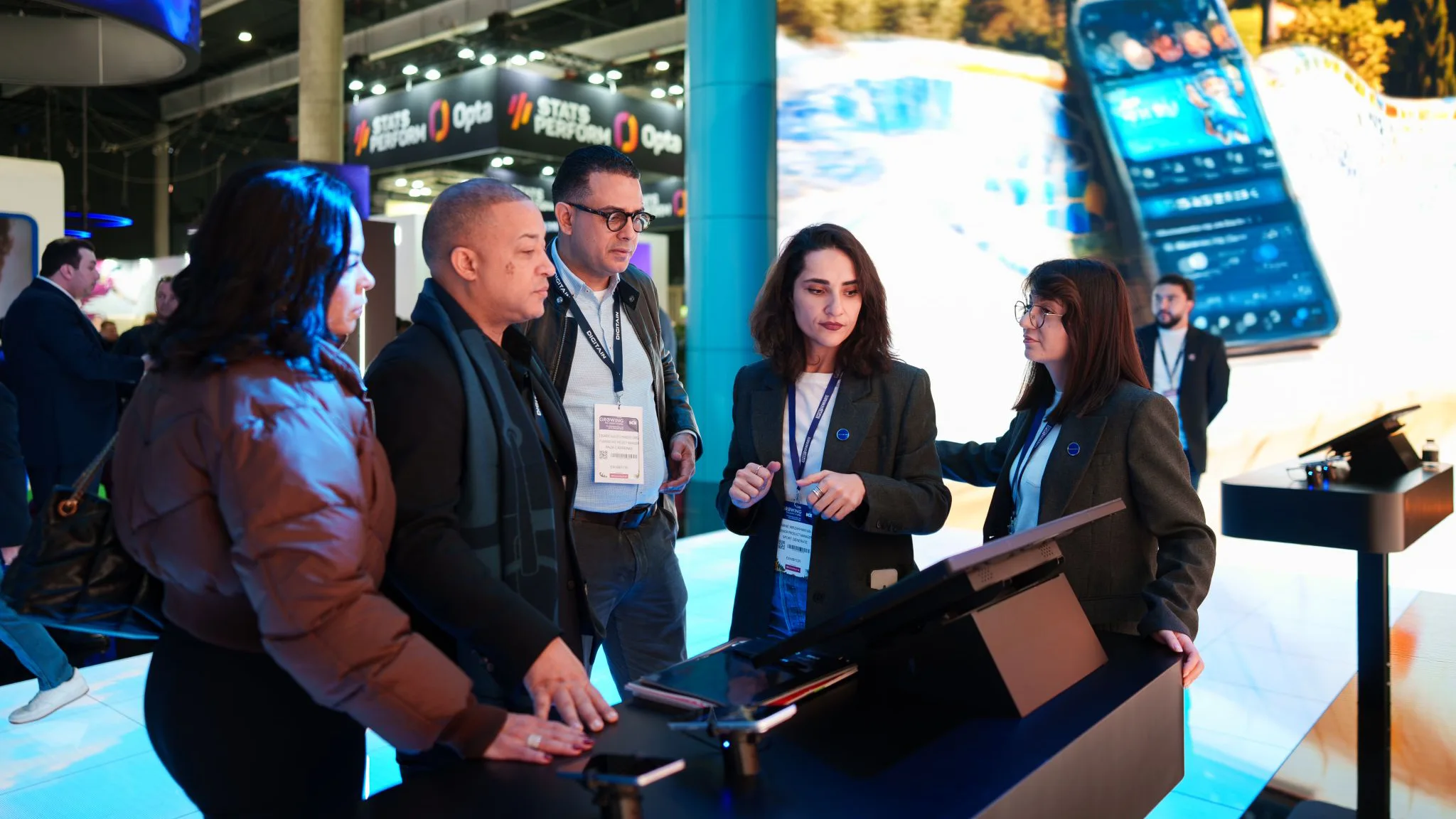 Digitain team demoing a touchscreen console with visitors gathered, bright mobile app visuals in the background.
