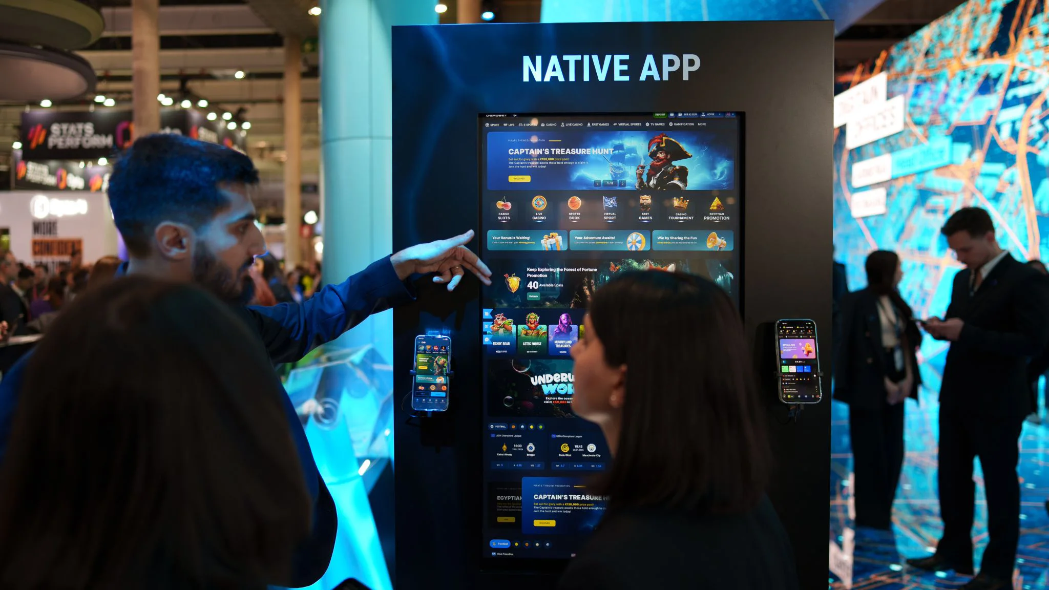 Presenter points at a tall “Native App” display showing the app interface while attendees watch at the stand.