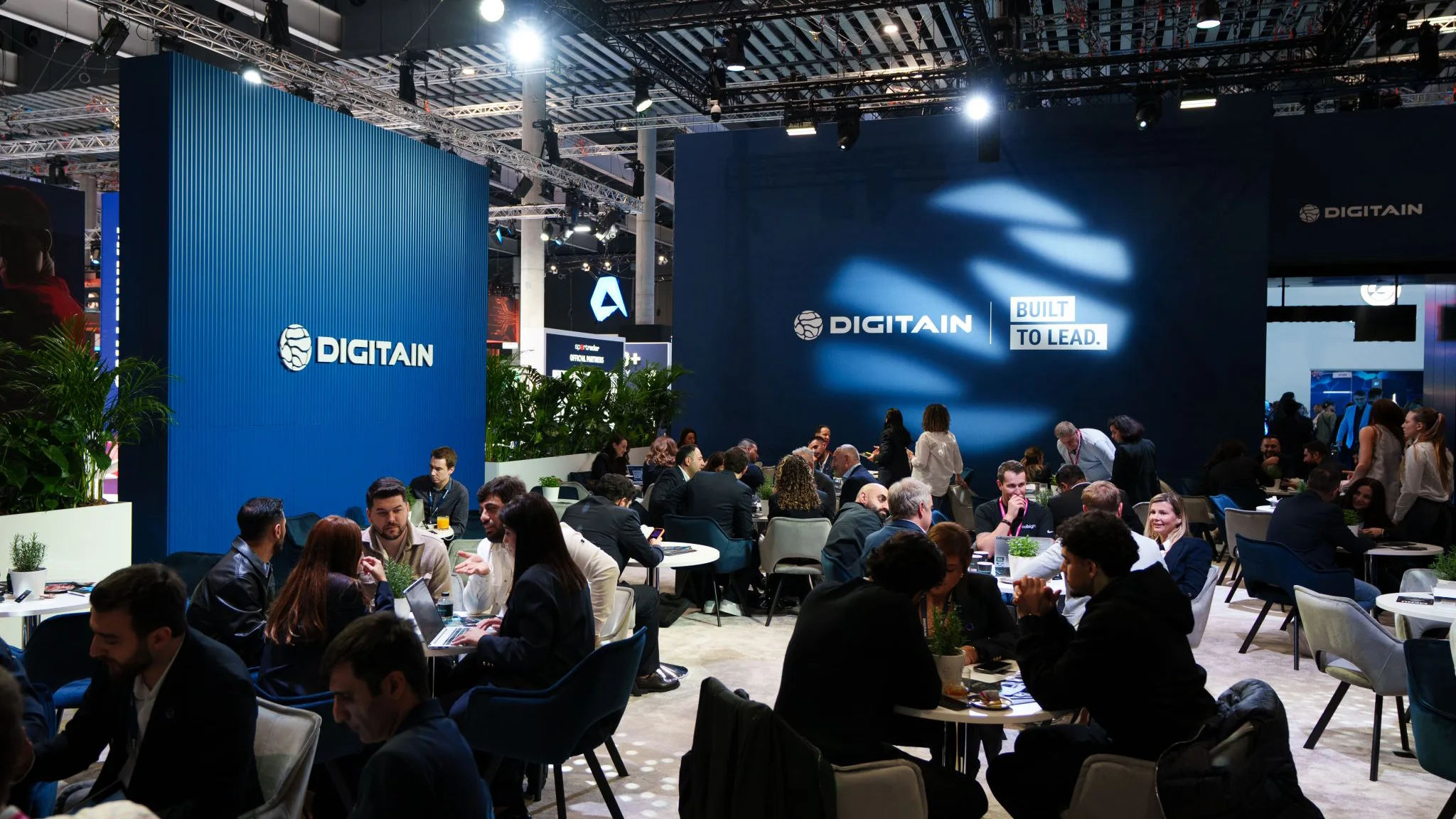 Digitain lounge area filled with seated meetings, large “Built to Lead” branding wall, and visible exhibition hall rigging.