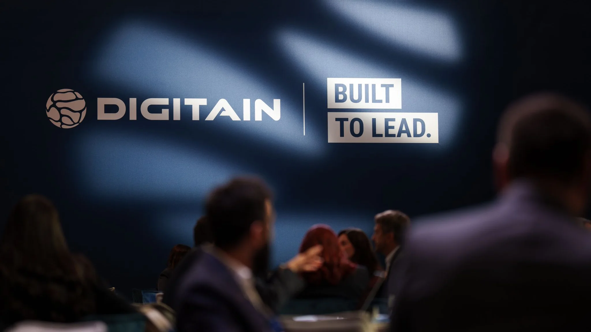 Digitain “Built to Lead” wall branding in focus, with visitors softly blurred in the foreground.