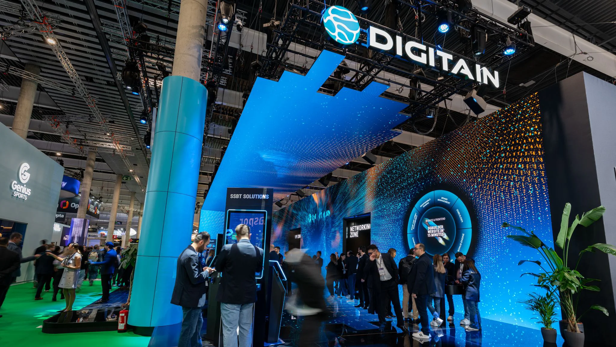 Wide angle of the Digitain stand with overhead logo, blue canopy lighting, and attendees entering the space.