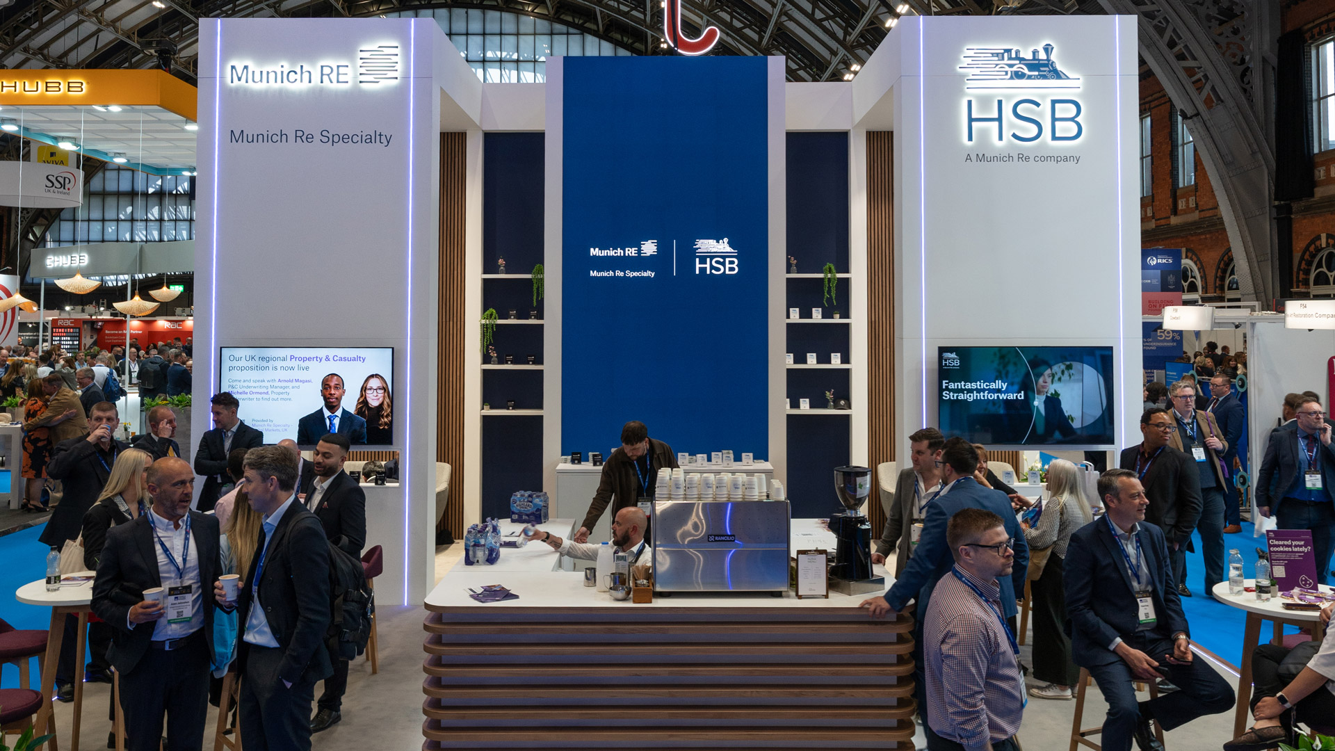 Front view of the Munich Re Specialty and HSB BIBA 2026 exhibition stand, showing the central hospitality bar, branded towers, digital screens and client meeting areas.