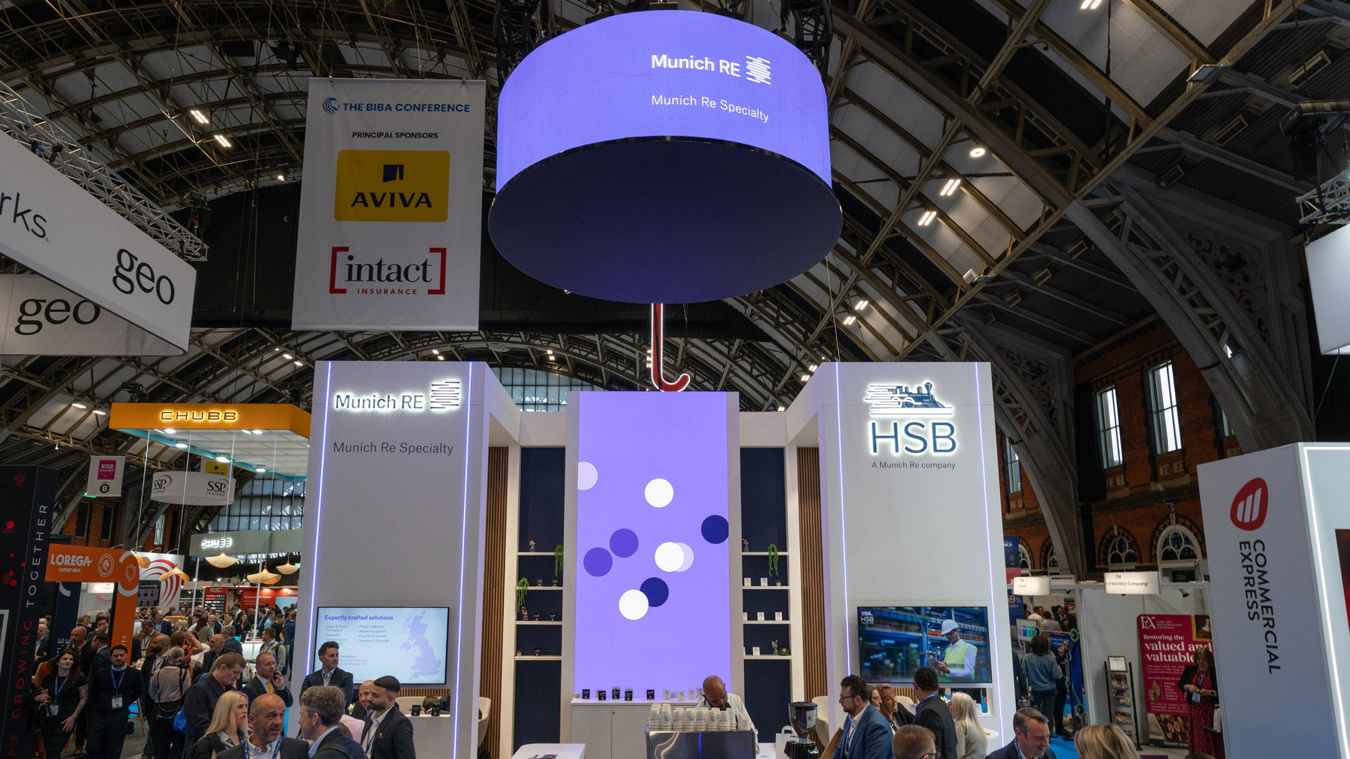 Munich Re Specialty and HSB stand at BIBA 2026 with overhead circular banner, illuminated brand towers, central LED feature wall and busy exhibition hall traffic.