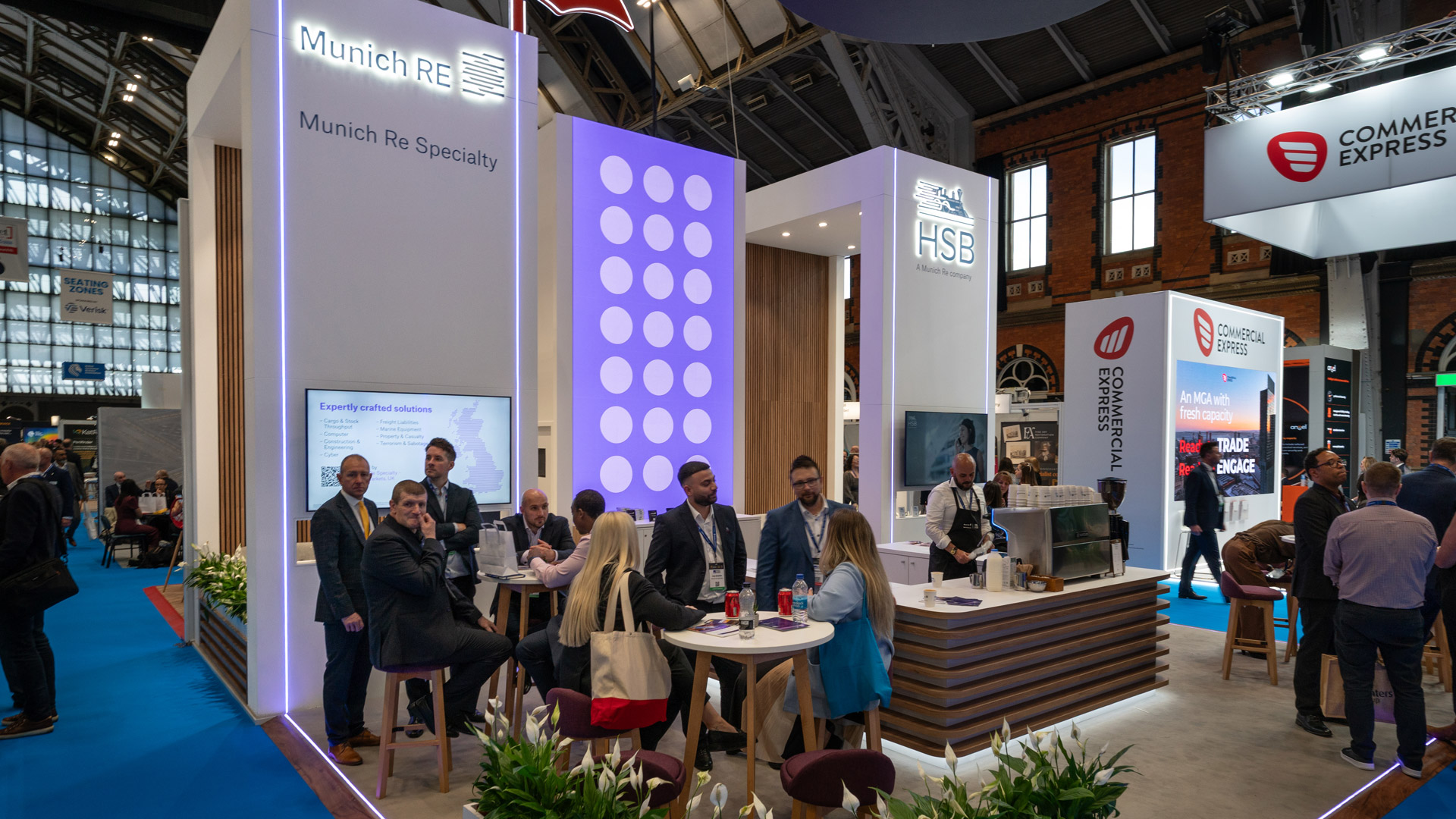 Munich Re Specialty and HSB BIBA 2026 exhibition stand with hospitality bar, semi private meeting tables, illuminated branding and central digital content wall.
